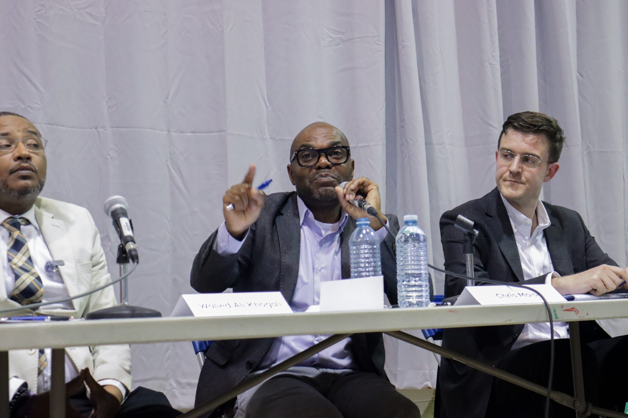 Chris Moise speaks into a microphone at a municipal debate, waving his finger at the audience as rival candidates watch him